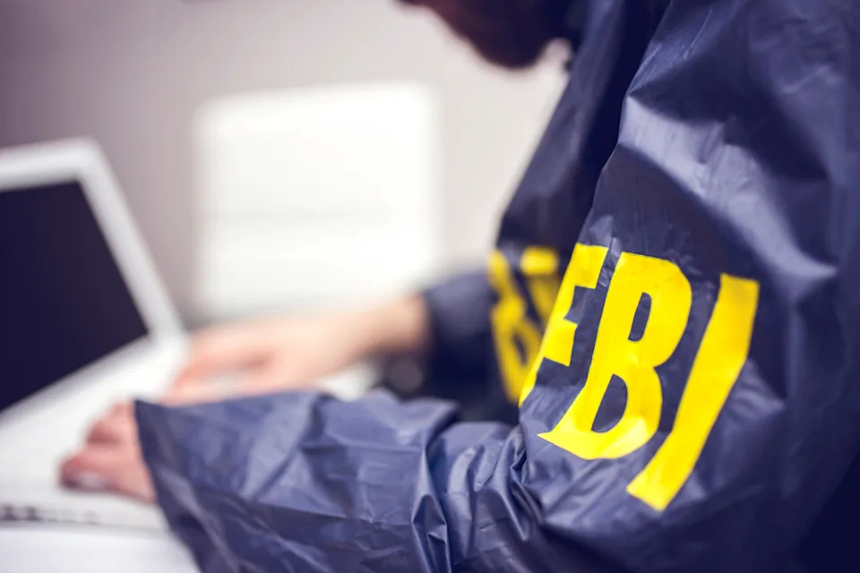 Person typing on a laptop, wearing a jacket labeled "FBI" on the sleeve