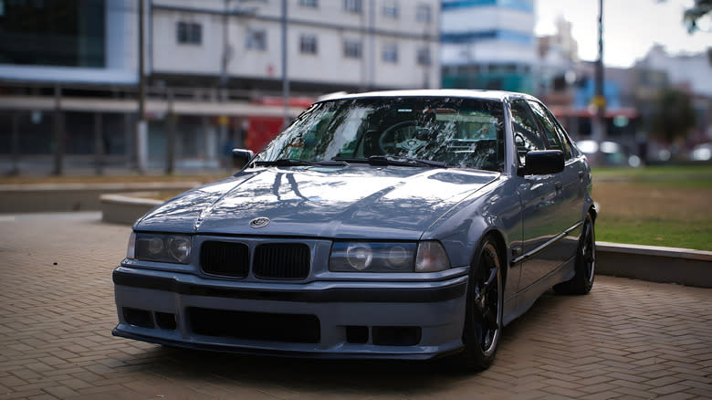 Juiz de Fora, MG, Brazil – July 12, 2025: Front-side view of a customized gray BMW E36 sedan parked in an open urban area, showcasing its sporty design, lowered suspension, black wheels.