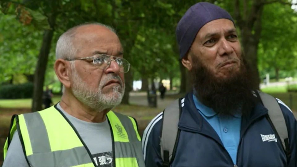 Two men stands next to each other. The one on the left is wearing a hi-vis jacket over a grey t-shirt and glasses, while the one on the right is wearing a blue polo and a blue tracksuit jacket. He also seems to be carrying a backpack.