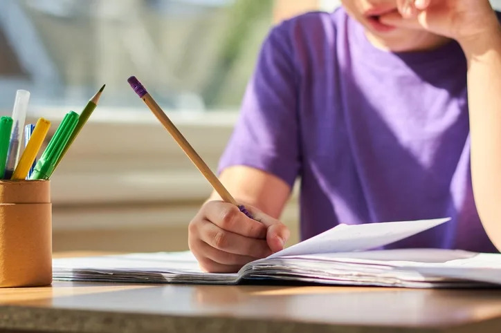 Child writing in a notebook at a desk with a pencil holder nearby, focusing intently on their work