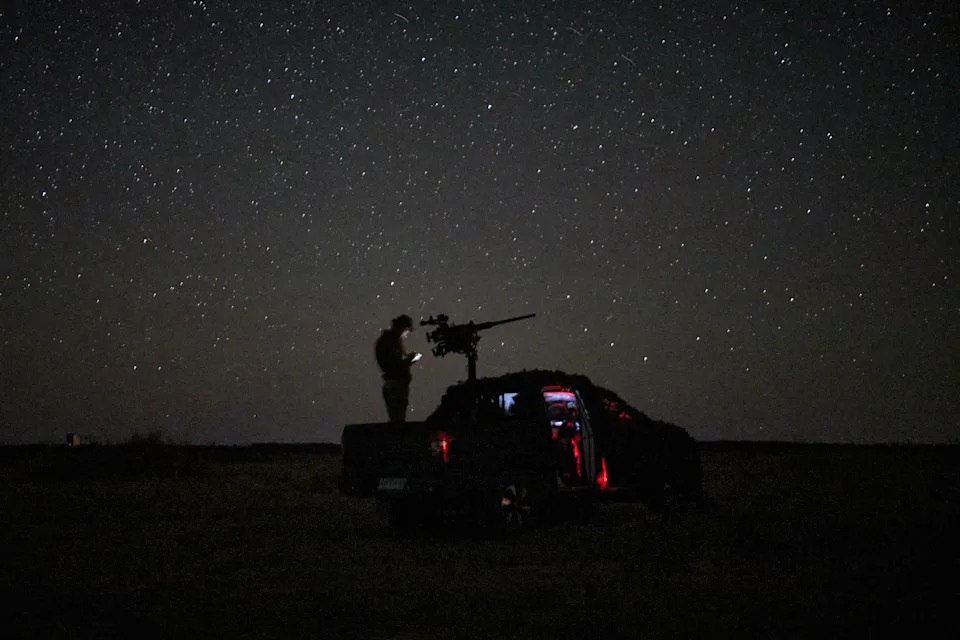 A soldier stands in a truck bed beside a mounted gun, seen under stars at night.