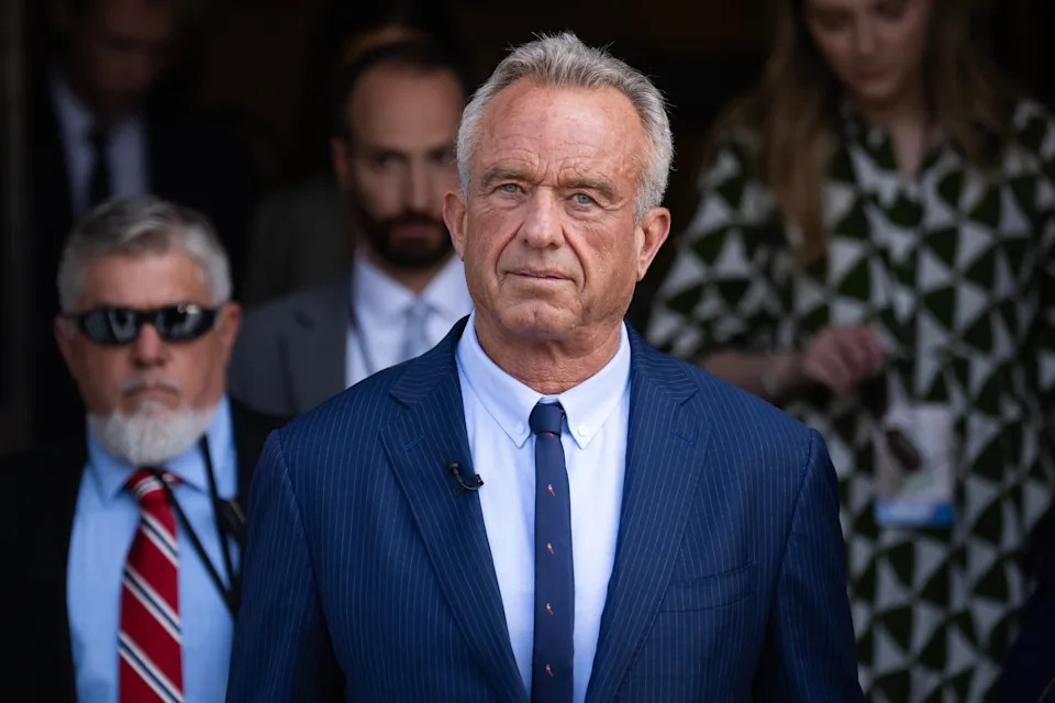 A man in a tailored suit with a polka dot tie appears serious, surrounded by individuals in a professional setting