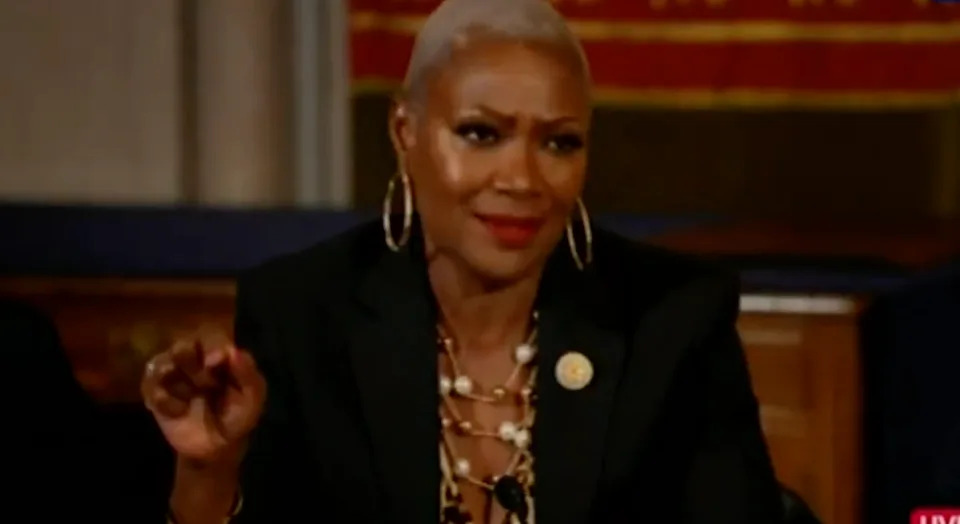 Person in a black suit with gold accents speaks animatedly, wearing hoop earrings and layered necklaces