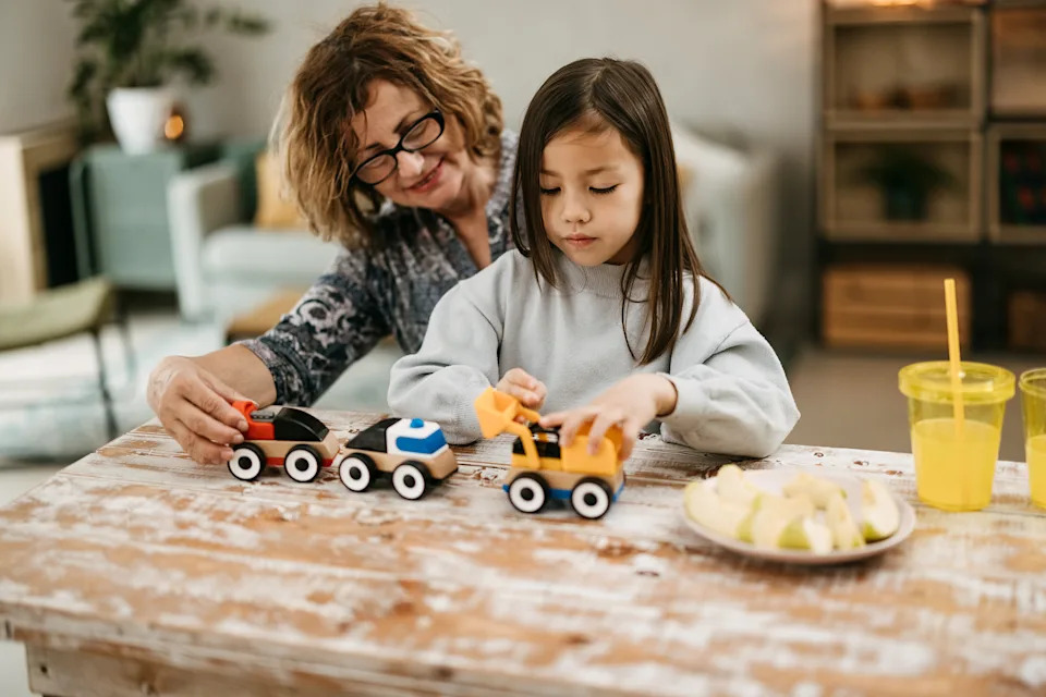 An older woman and a young girl play with wooden toy trains at a table, with apple slices and drinks nearby, creating a nurturing moment