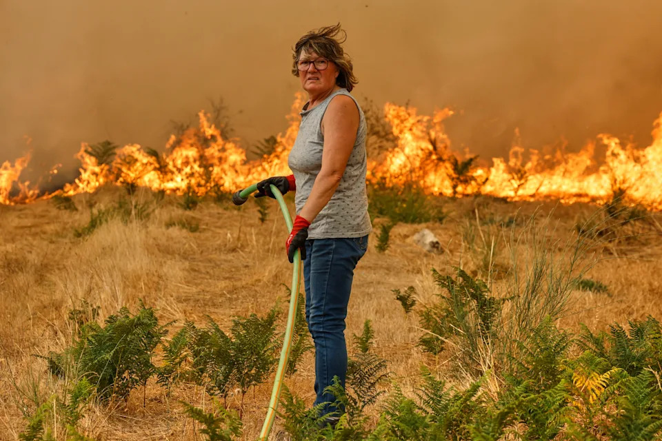 A person holds a hose while standing in a field in front of an oncoming wildfire.