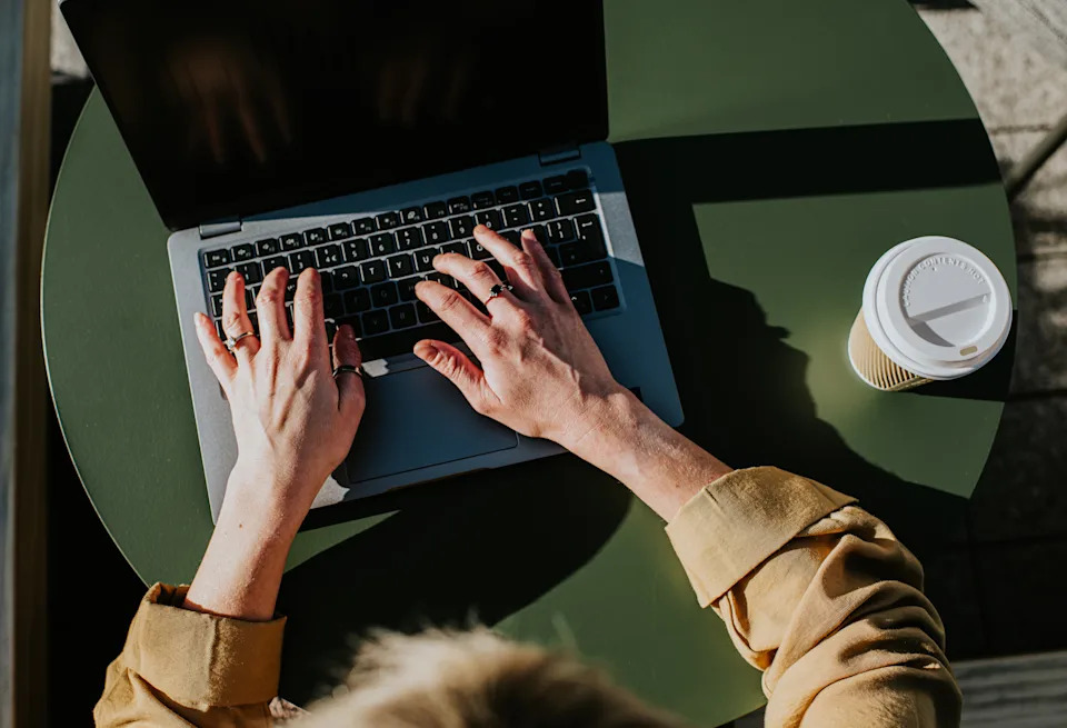 Person typing on a laptop at an outdoor table with a cup of coffee beside them