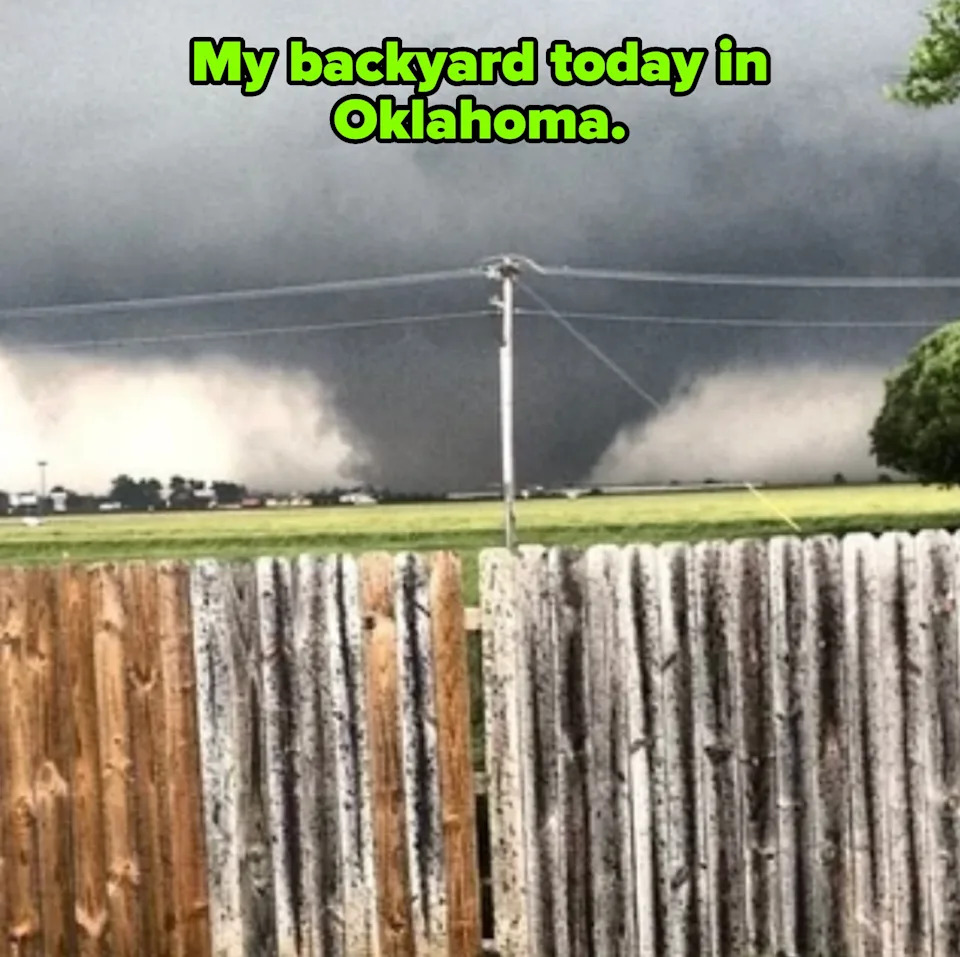 Tornado in the distance beyond a wooden fence, with dark storm clouds overhead and fields in the foreground