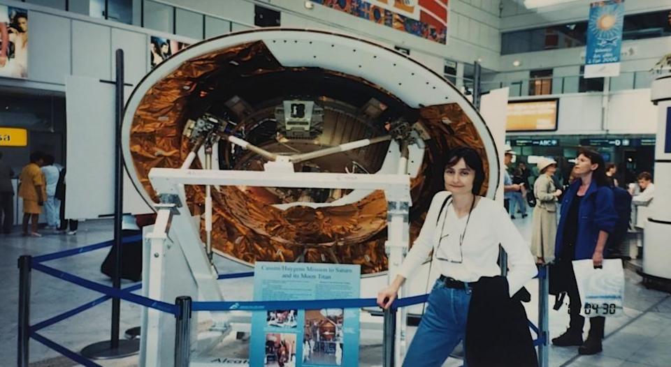 Michele Dougherty, de 30 años, con un top blanco y vaqueros azules, gafas colgando del cuello y una chaqueta negra colgada del brazo. Está de pie delante de una parte redonda de una nave espacial, en parte blanca pero con partes de lámina de cobre. Está delimitada por una barandilla azul. La escena parece transcurrir en un museo, o tal vez en un aeropuerto, y hay gente alrededor.