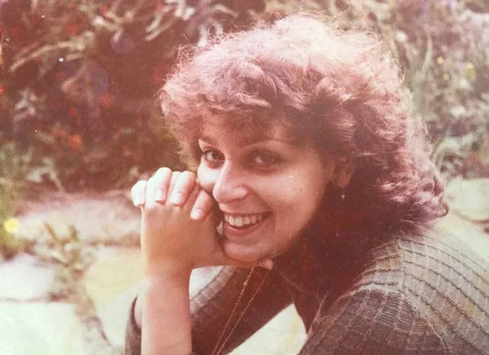 Person smiling outdoors with curly hair, wearing a textured, long-sleeved top, resting hands near face