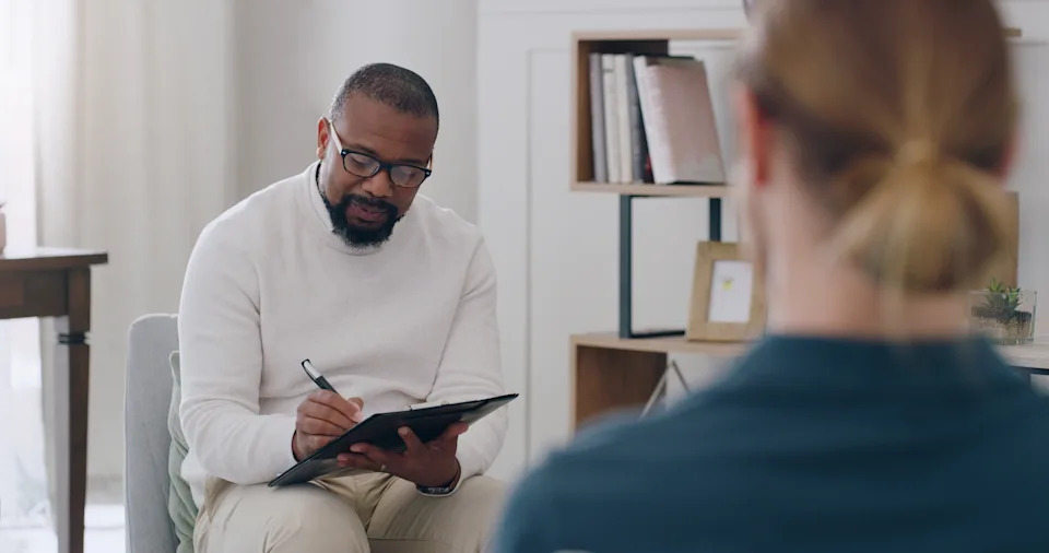 A therapist sits and writes notes on a clipboard while attentively listening to a person seated across from them