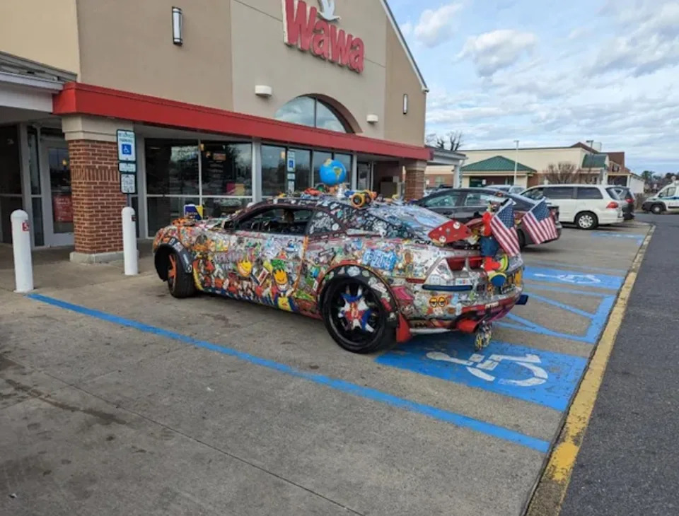 Car parked in a handicap spot, covered in various colorful stickers and decorations, with American flags on the rear, outside a Wawa store