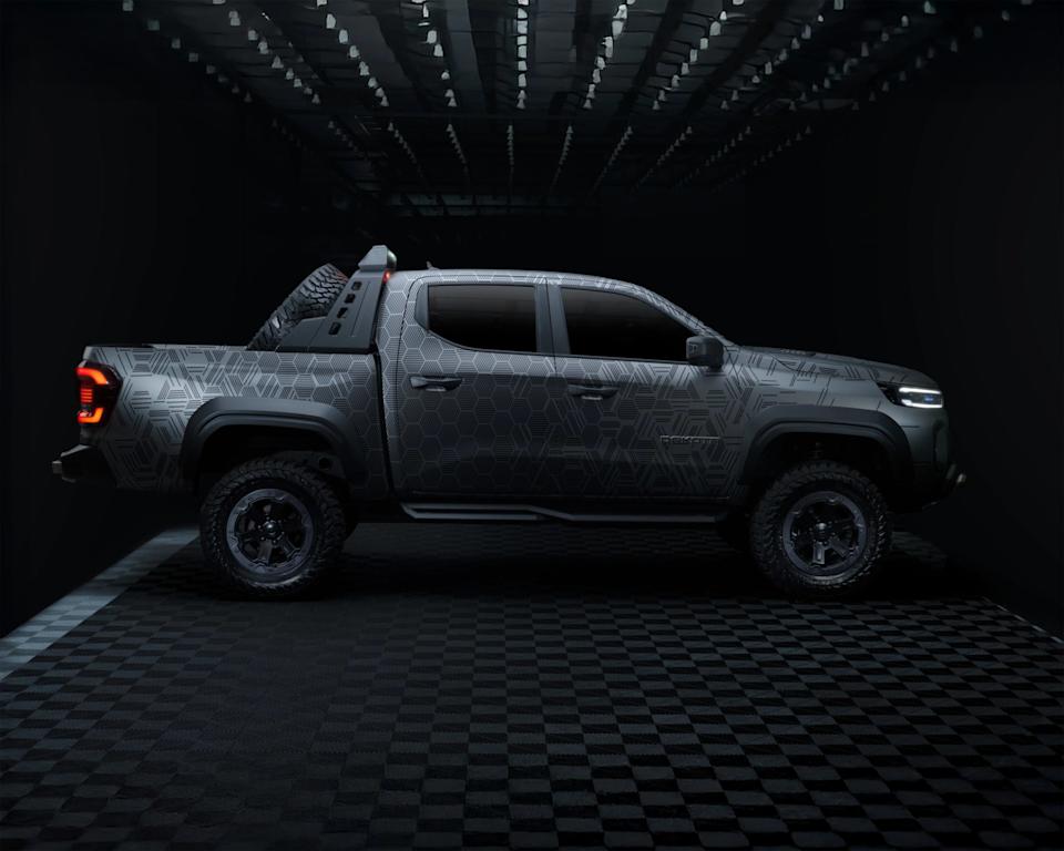 Side view of a modern pickup truck with a geometric patterned matte gray finish, large off-road tires, and a spare tire mounted in the bed. The truck has tinted windows, black fender flares, and sleek LED headlights and taillights. The background is dark with a checkered floor and overhead lighting.