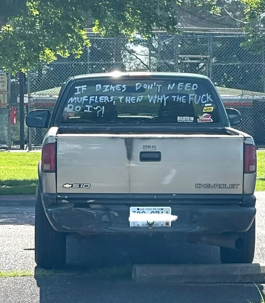 Truck with hand-painted sign on rear window: "If bikes don't need mufflers, then why the [expletive] do I?"