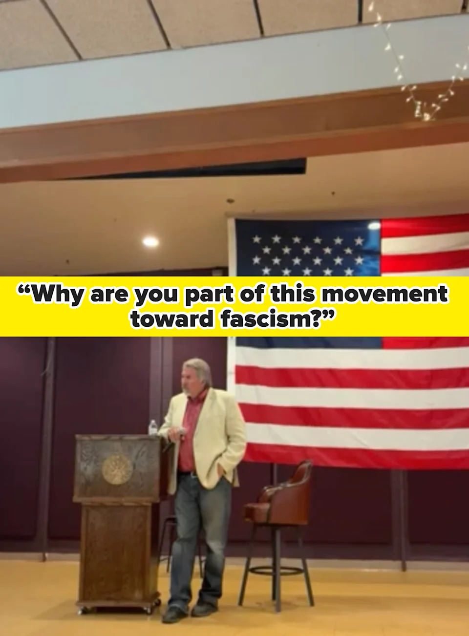 A person in a blazer stands at a podium with a microphone, next to a large American flag