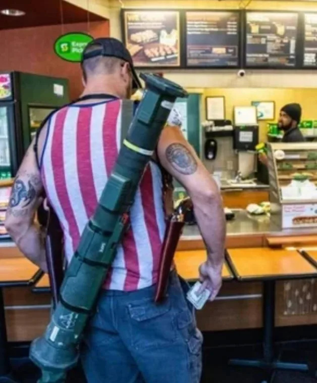 Man in a sleeveless shirt with stars and stripes at a sandwich shop counter, holding cash. He has a rocket launcher strapped to his back