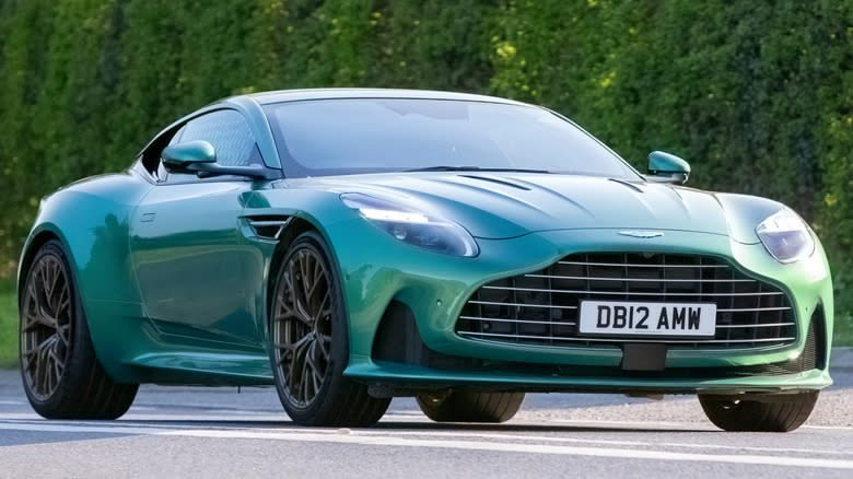 A green Aston Martin DB12 sports car drives in front of dense foliage