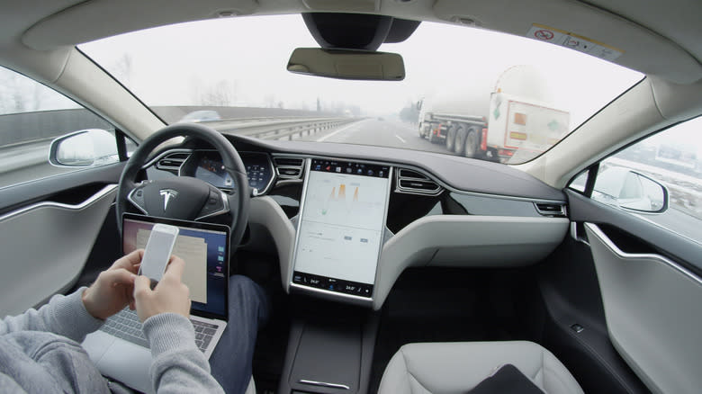 Self-driving Tesla Model S car autopilot demanding driver attention to hold steering wheel take control on highway. Man working on laptop texting on mobile