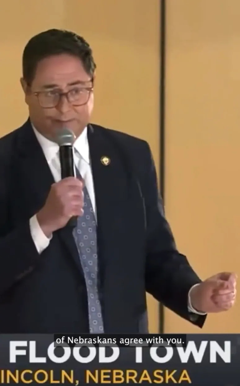 A person in a suit speaks into a microphone at an event labeled "Flood Town, Lincoln, Nebraska."