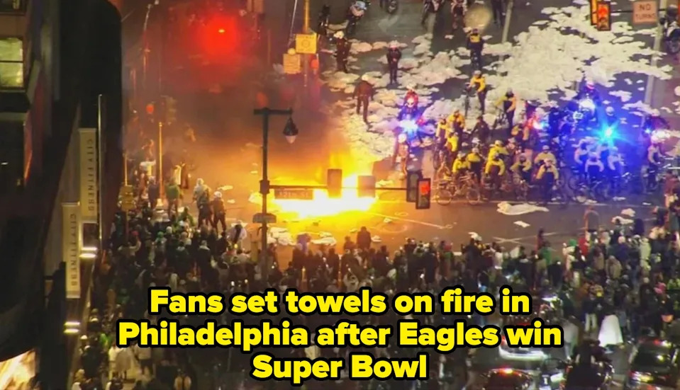 Aerial view of a chaotic street scene with a fire, crowd, and police presence creating a tense atmosphere at night. Caption: "Fans set towels on fire in Philadelphia after Eagles win Super Bowl"