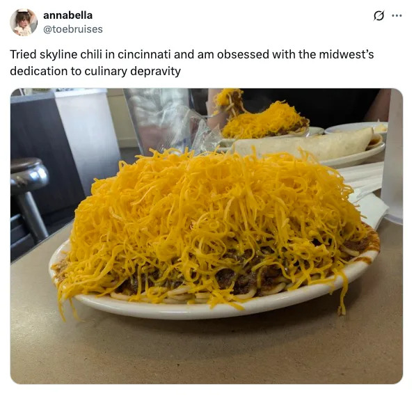 A plate piled high with shredded cheese atop Skyline chili, with a person's hands and a table visible in the background