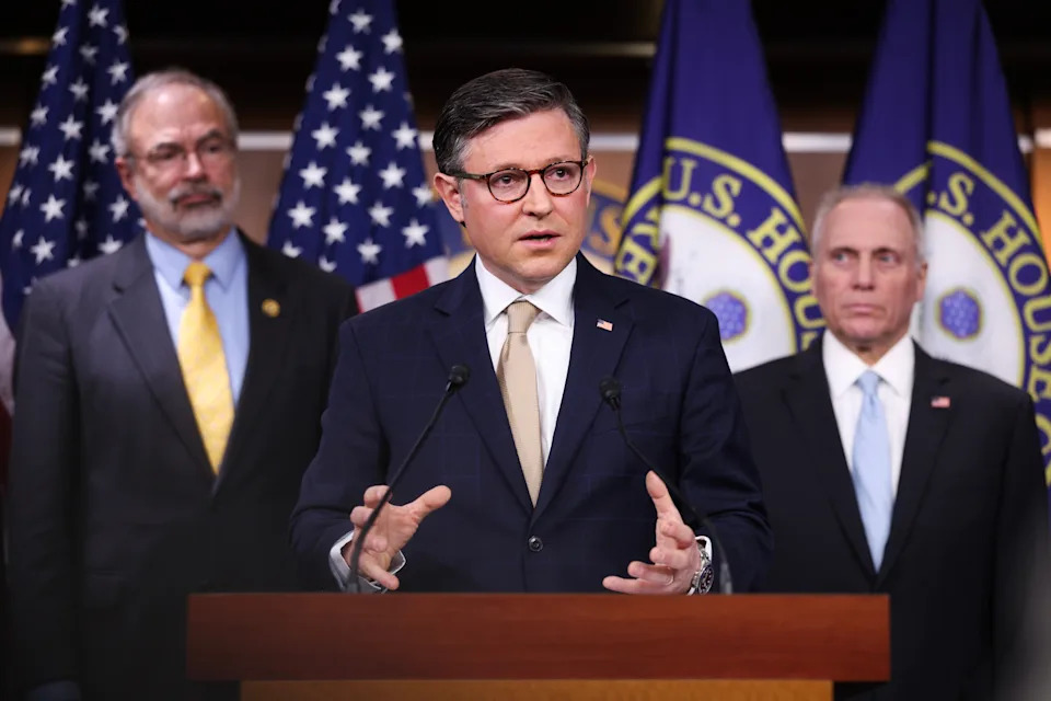 A man in a suit speaks at a podium with U.S. House seals and flags behind, flanked by two others in suits, suggesting a formal announcement