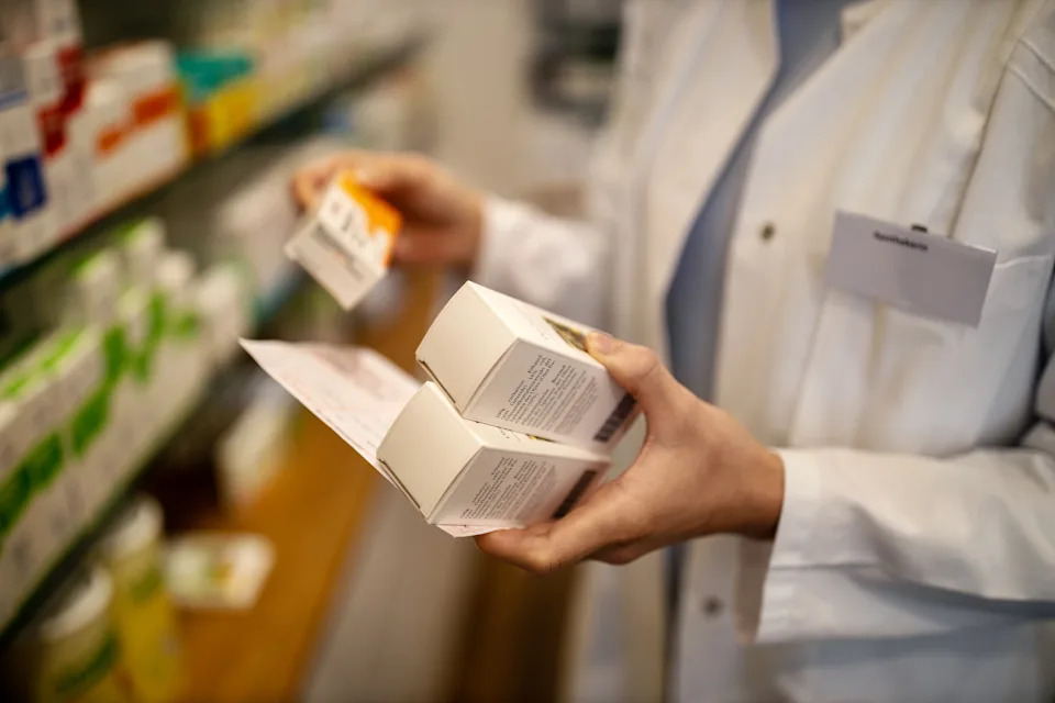 Pharmacist in a lab coat examines medication boxes and a pamphlet in a pharmacy setting