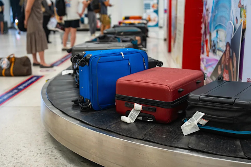 Luggage rotates on an airport baggage carousel, with travelers in the background picking up their bags