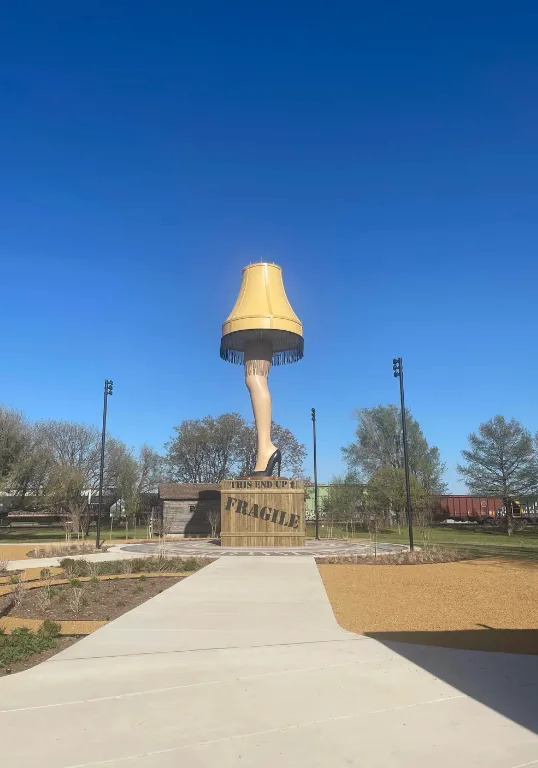 Statue of a large lamp resembling a leg with a shade, similar to the iconic lamp from "A Christmas Story," set in an outdoor park