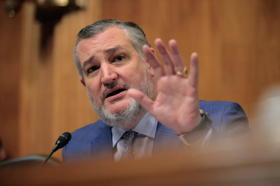 A person gestures while speaking at a microphone, wearing a suit and tie, during a formal event or meeting
