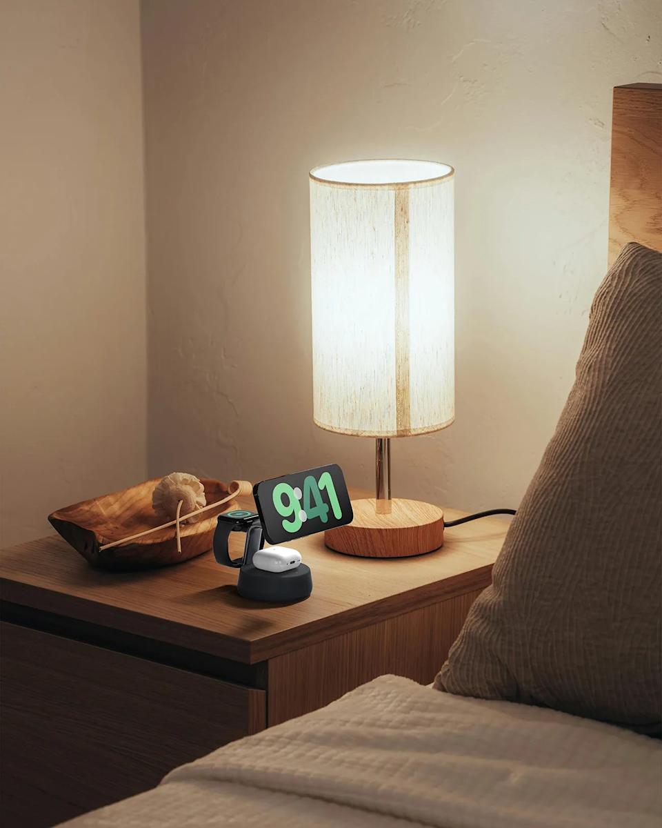 Wooden bedside table with a modern cylindrical lamp featuring a light beige fabric shade. On the table, there is a black multi-device charging stand holding a smartphone displaying the time 9:41, a smartwatch, and wireless earbuds. A wooden decorative bowl with a natural scrub brush is also on the table. The bed next to the table has a beige textured pillow and a light-colored quilted blanket.