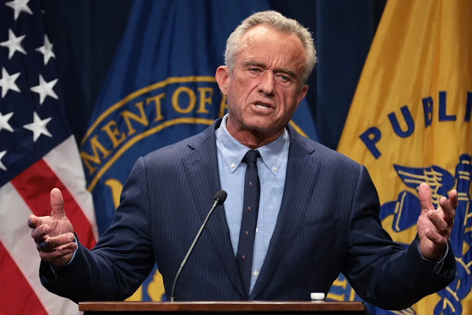 A person in a suit speaks at a podium, gesturing with both hands, in front of official-looking banners