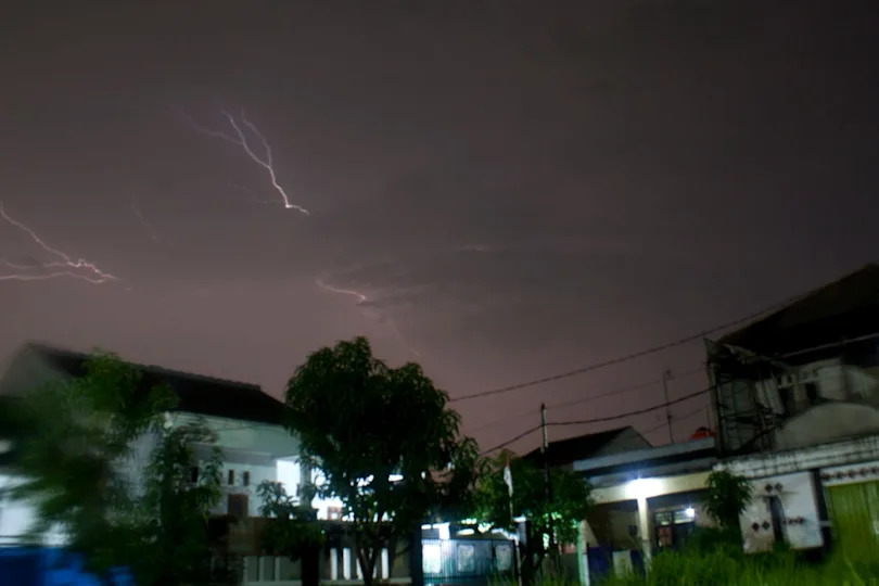 Lightning streaks across the night sky over a quiet residential neighborhood with several houses silhouetted in the foreground