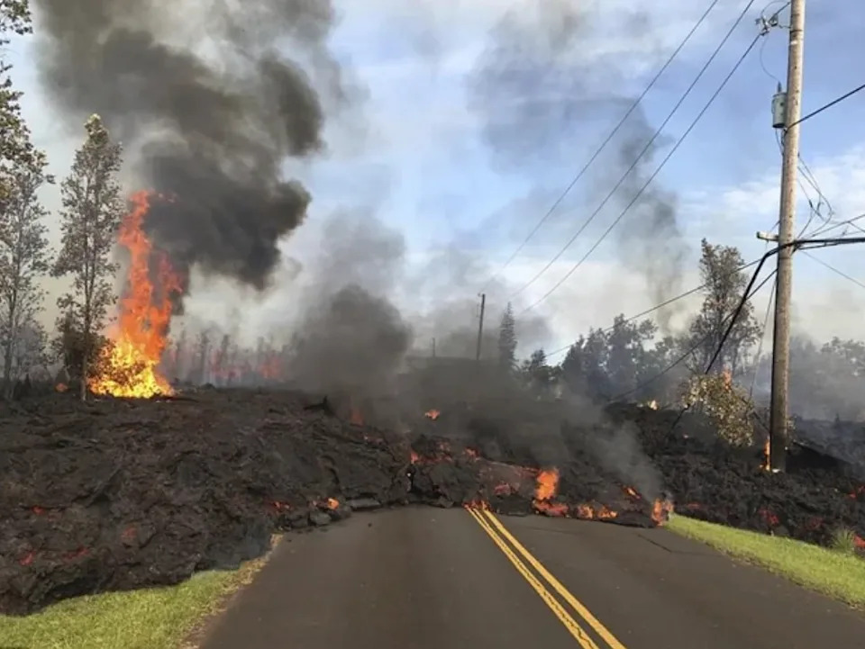 Lava flows over a road, with flames and smoke rising, surrounded by trees and utility poles