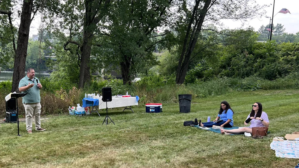 A church sermon being held in an outdoors space with people sitting in the grass.