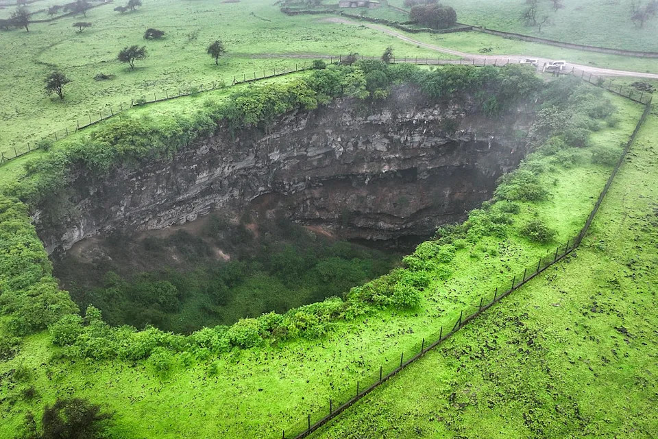 Aerial view of a large sinkhole surrounded by green grass and some trees, with a fence encircling the perimeter