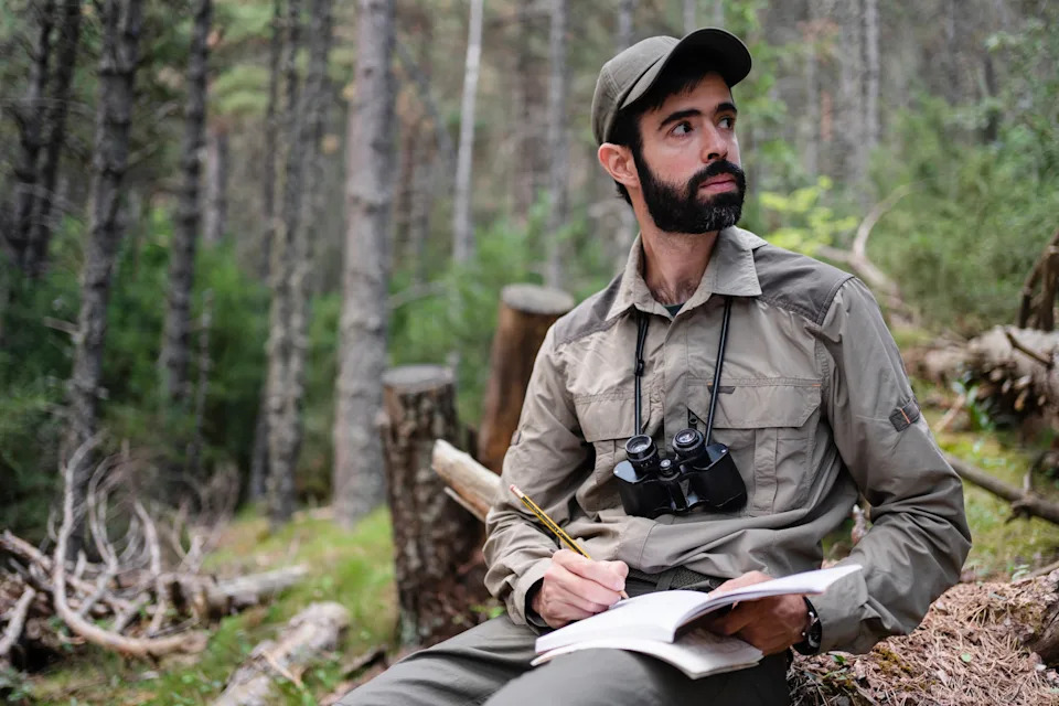 Person in a forest setting wears outdoor gear and binoculars, writing in a notebook, appearing focused and contemplative