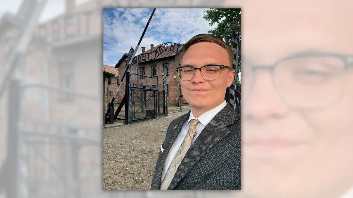 A white man stands in front of an older building with a gate in front wearing a gray suit. 