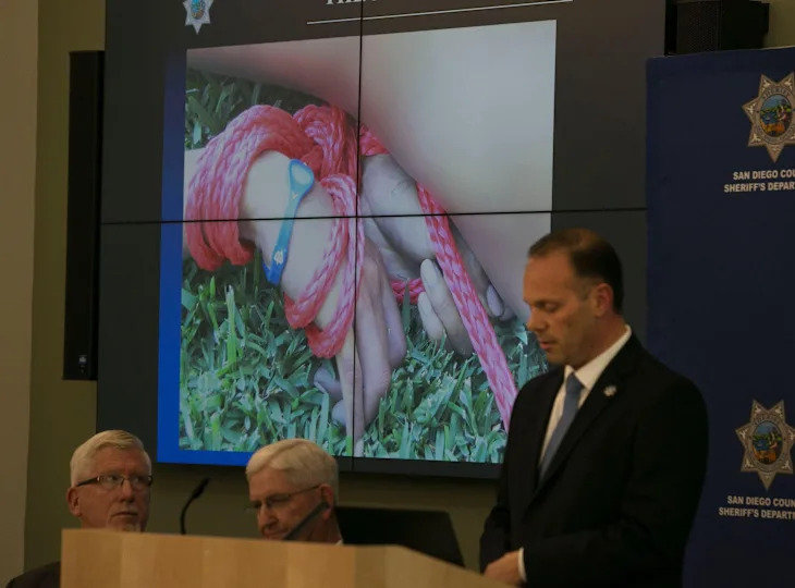 A press conference with a speaker at a podium and a screen displaying an image of hands tied with rope