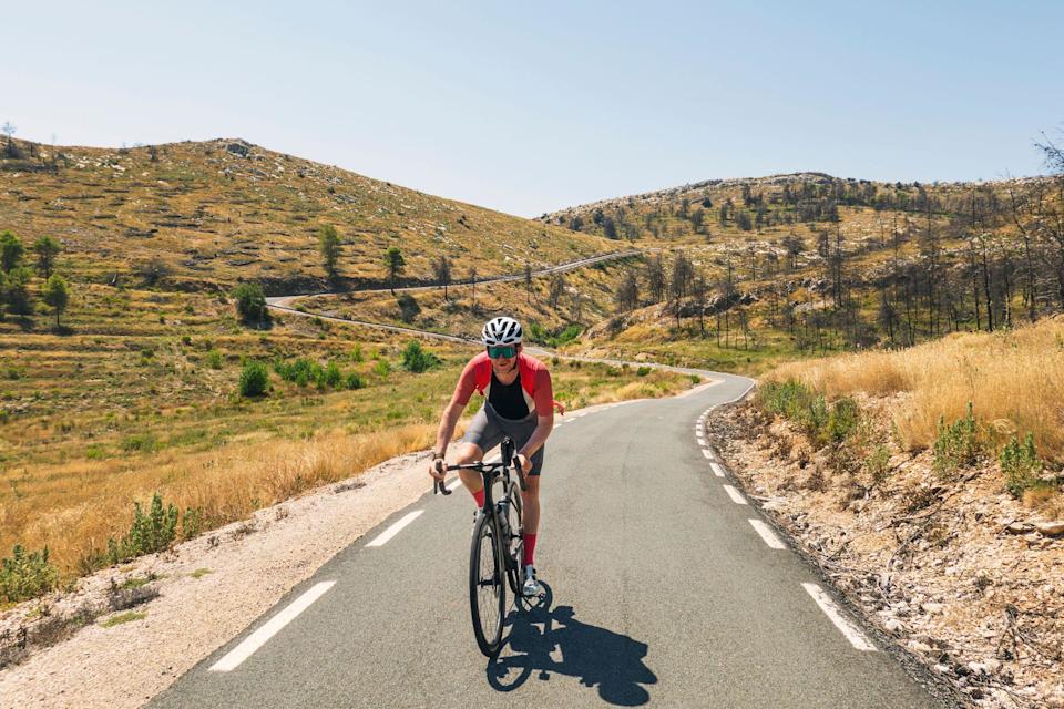 Una persona sobre una bicicleta en un soleado día de verano desciende una serpenteante carretera que baja de dos altos montes.