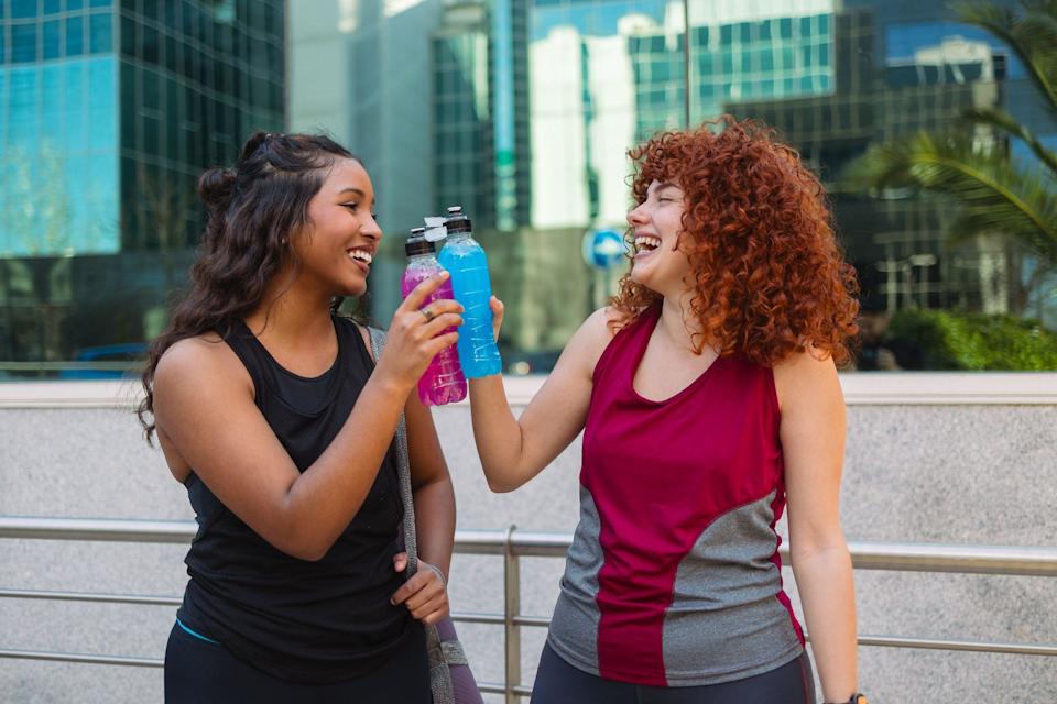 Dos mujeres en atuendo de ejercicio, se miran riendo y hacen brindis con dos bebidas isotónicas.