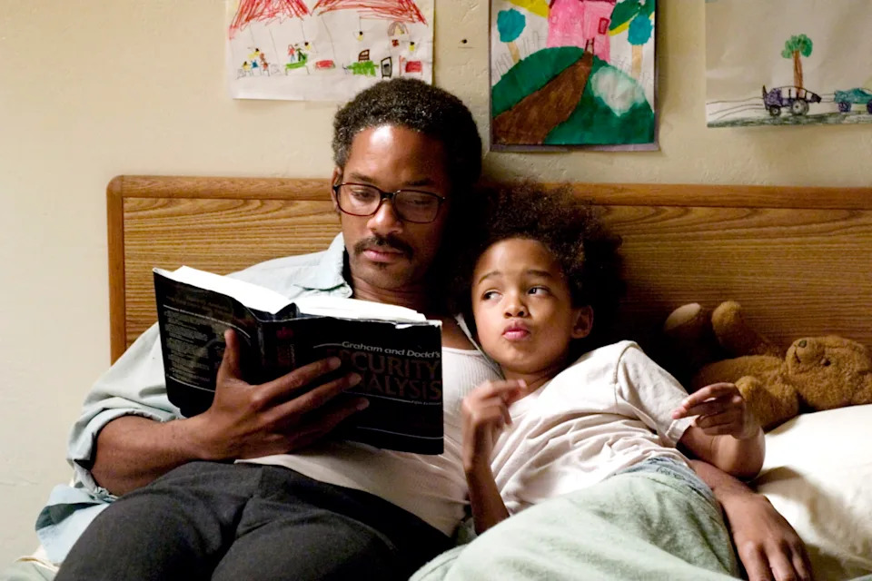 A person reads a book beside a child in bed, with drawings on the wall and a teddy bear nearby