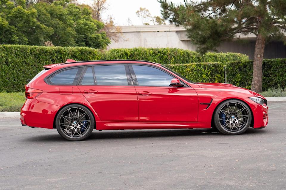 side view of red station wagon with black wheels, roof rails, and flared fenders parked near tree.