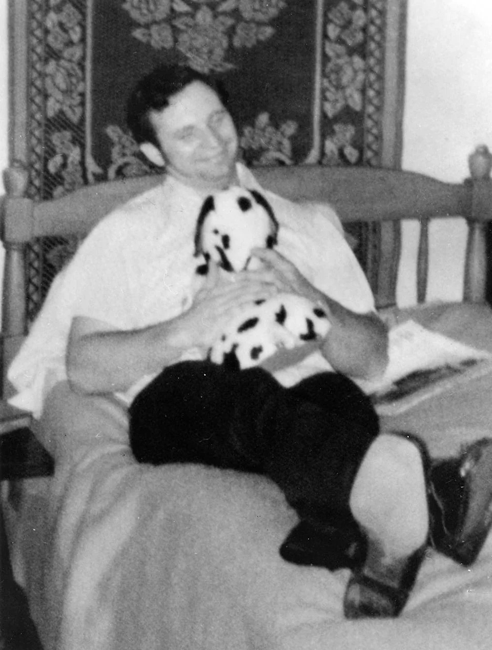 Dean Corll laying on a bed holding a stuffed animal smiling.