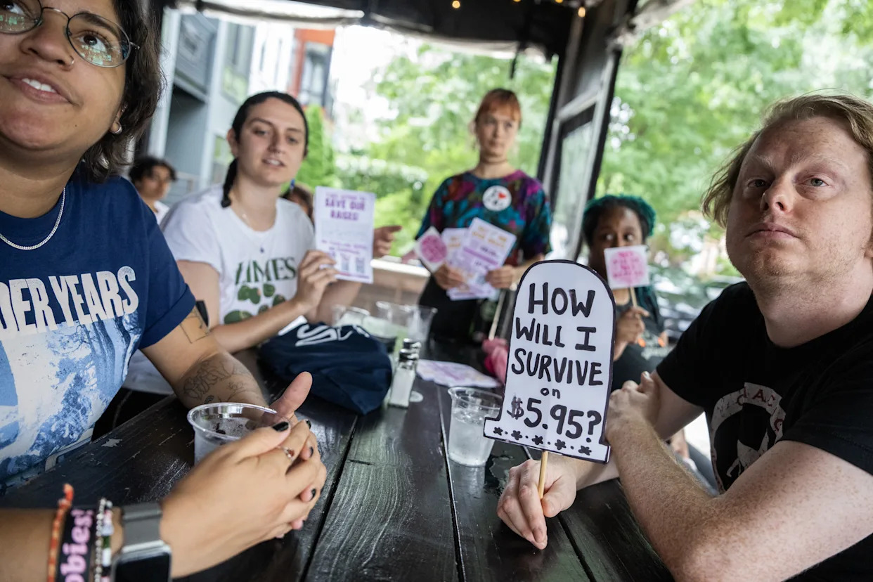 Five people sit around a table with a handwritten sign that says: &#x00201c;How will I survive on $5.95?&#x00201d;