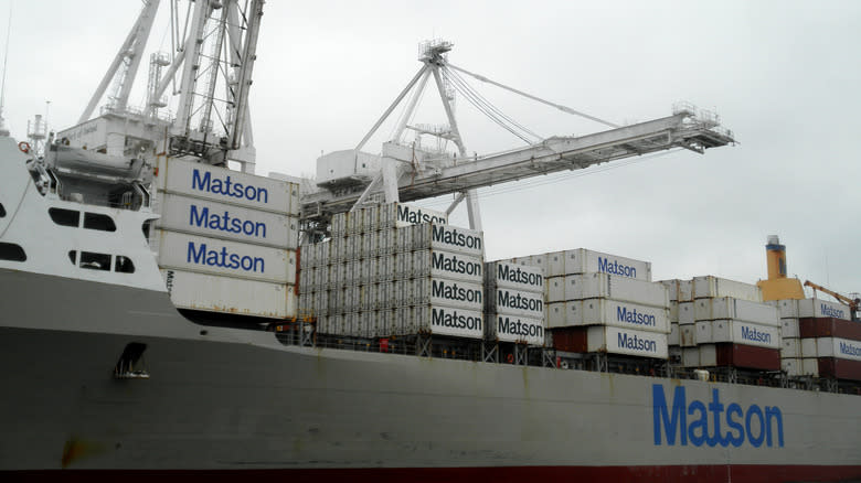 Matson Cargo Ship Mahi Mahi loaded with goods at Port by cranes in Oakland Harbor as a tugboat pushes a barge in front of it.