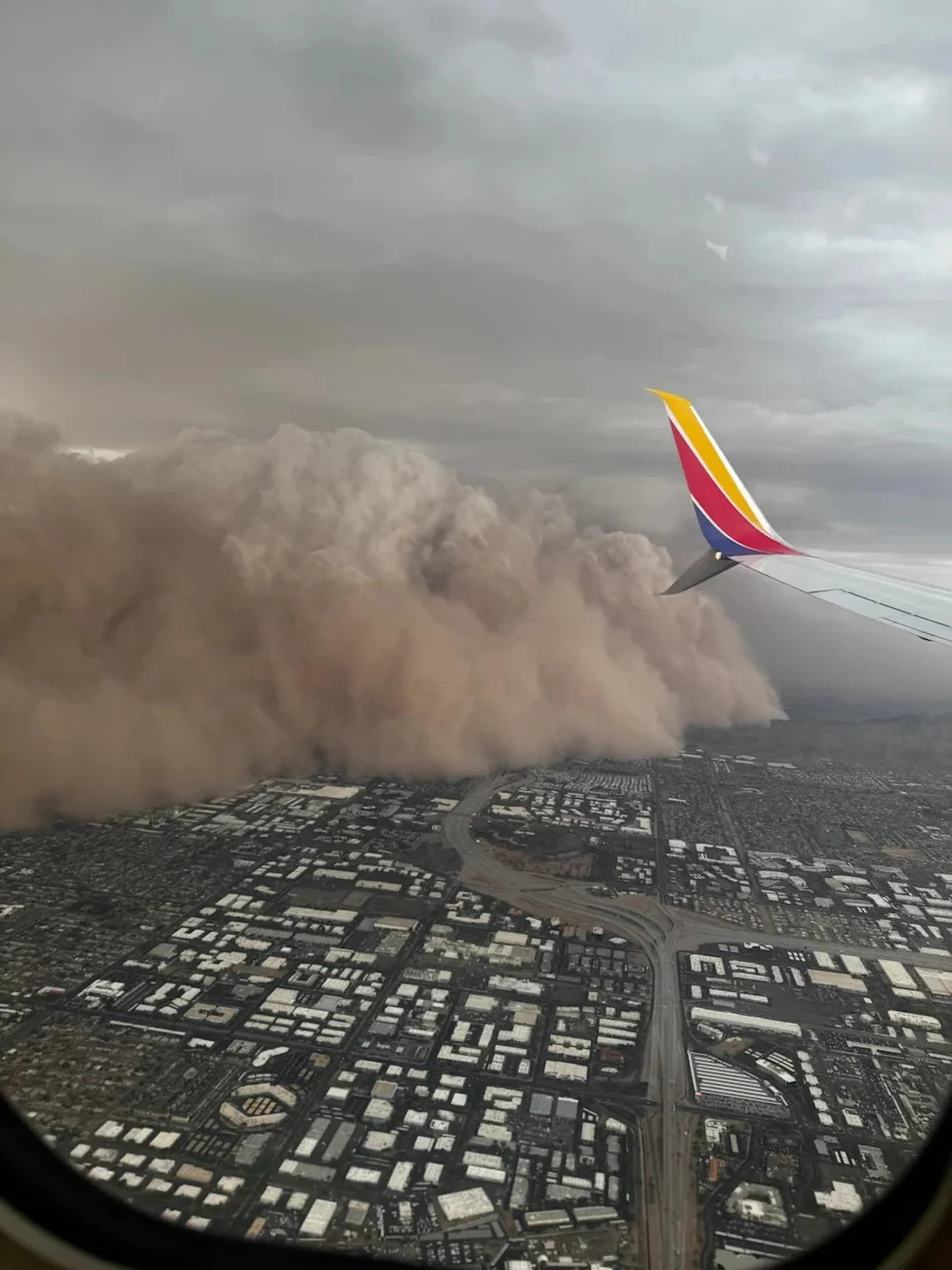 View from a plane window of a massive dust storm approaching a city, highlighting the stark contrast between urban landscape and nature