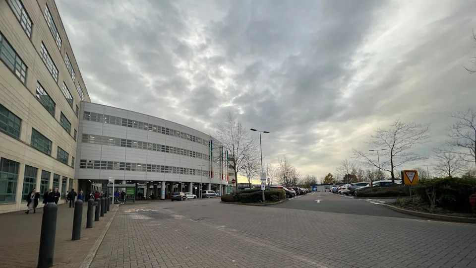 A view of a hospital building with a car park to the right side. It is a cloudy day.