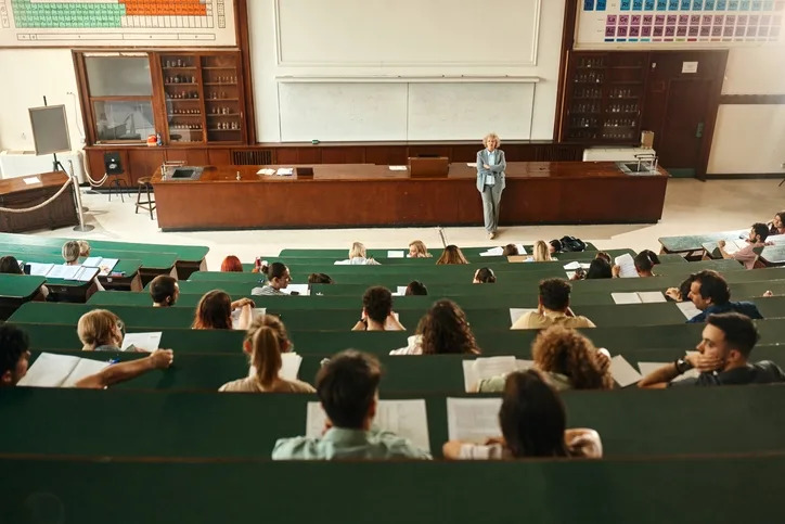 Large lecture hall with many students seated in tiered rows, listening to a professor standing at the front with a periodic table on display