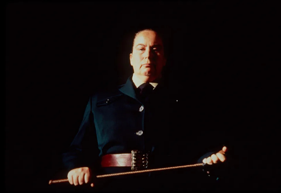 Person in formal attire holding a riding crop, standing against a dark background