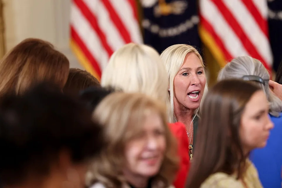 A woman speaks passionately in a crowded room. People around her are engaged and listening. Flags are visible in the background